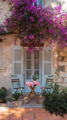 two chairs and a table in front of a building with purple flowers growing over it