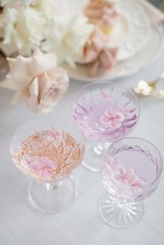 three wine glasses sitting on top of a table next to white and pink flowers in vases