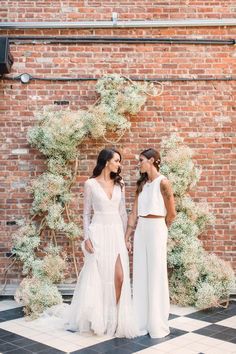 two women standing next to each other in front of a brick wall with greenery