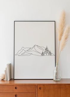 a vase with some flowers on top of a wooden dresser next to a framed drawing