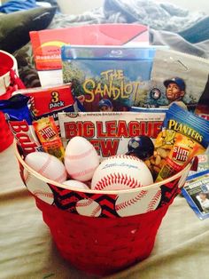 a red basket filled with baseballs and other sports related items sitting on top of a bed