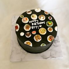 a black birthday cake with lots of food on the top and writing happy birthday byyi