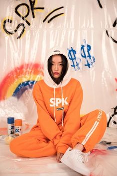 a woman sitting on the ground wearing an orange sweat suit and white hoodie, with spray paint all over her face