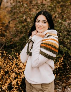 a woman standing in front of some bushes wearing a striped knitted sweater and scarf