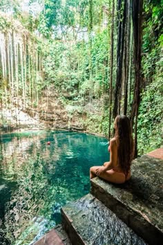 a woman sitting on the edge of a body of water in front of a forest