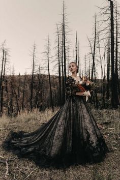 a woman in a long black dress standing in front of burned trees and holding flowers