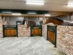 two horses are standing in an enclosed area with brick walls and green gated stalls