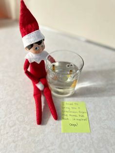 an elf is sitting on the table next to a glass of water and a note