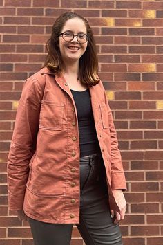 a woman standing in front of a brick wall wearing black pants and an orange jacket