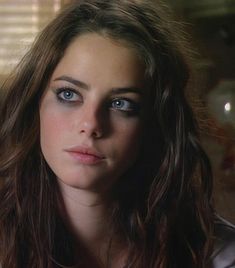 a woman with long hair and blue eyes looks into the camera while standing in front of a window