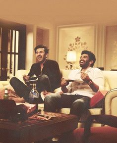 two men sitting on couches laughing and drinking wine in a living room with other people