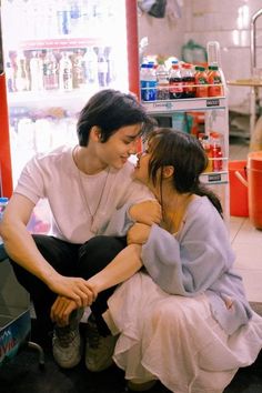 two people sitting on the ground in front of a vending machine and one is kissing the other's cheek