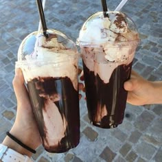 two people holding up drinks with ice cream and chocolate toppings on them in front of a cobblestone walkway