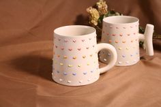 two coffee mugs sitting on top of a brown cloth covered table next to flowers