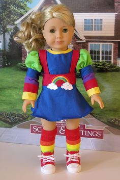 a doll with blonde hair wearing a blue dress and rainbow socks stands in front of a real house