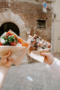 two slices of pizza are being held up in front of a brick building on the street