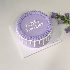 a purple birthday cake with white icing and flowers on the table next to it