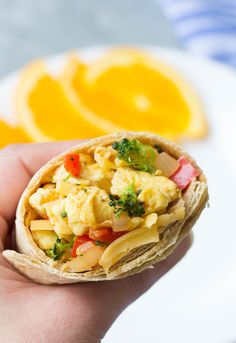 a hand holding a tortilla filled with food and veggies next to an orange slice