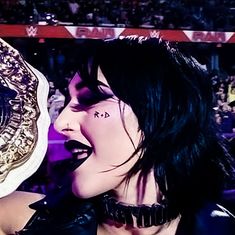 a woman with black hair and piercings holding up a wrestling belt in front of an audience