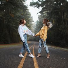 two young women holding hands while walking down the middle of an empty road with trees in the background