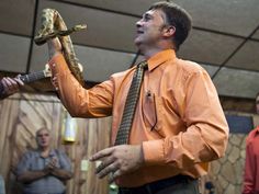 a man in an orange shirt and tie holds a snake on his neck while another man plays guitar behind him