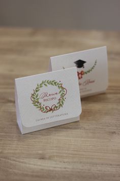 two business cards on a wooden table with a graduation cap and wreath design in the middle