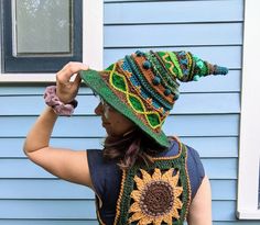 a woman wearing a crocheted sunflower hat on her head, standing in front of a blue house