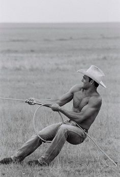 a shirtless man with a lasso in his hand sitting on the ground next to an open field