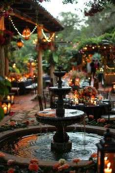 a water fountain surrounded by flowers and lite up lanterns in the background with people sitting at tables around it