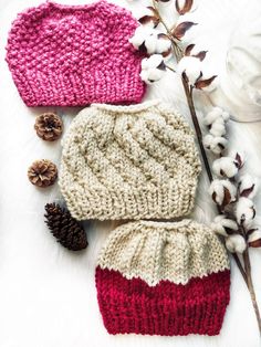 two knitted hats sitting on top of a white blanket next to cotton flowers and pine cones