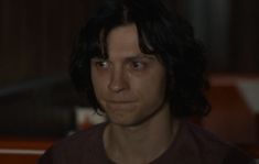 a man with long black hair wearing a brown shirt and looking at the camera in a dark room