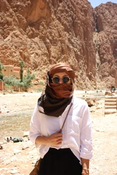 a woman wearing sunglasses and a scarf standing in front of a rocky cliff face with her hands on her hips