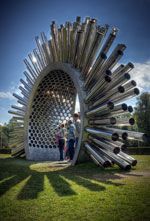 two people standing in front of a sculpture made of metal pipes and tubes on top of grass