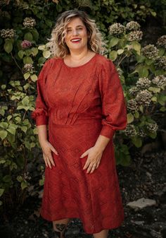 a woman wearing a red dress standing in front of some bushes and flowers with her hands on her hips