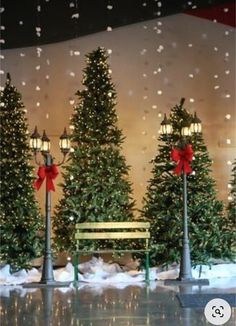 christmas trees in front of a building with snow falling on the ground and benches under them