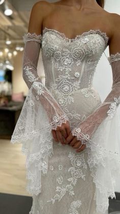 a woman in a white wedding dress with long sleeves and flowers on the skirt is holding her hands together