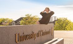 a woman sitting on top of a bench in front of a university sign with trees and buildings behind her