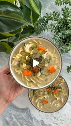 a hand holding a bowl of soup with carrots and noodles in it on a marble surface