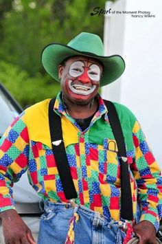 a man in clown makeup and suspenders is standing next to a white truck with his mouth open