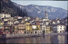 an old town is next to the water with mountains in the back ground and trees on both sides