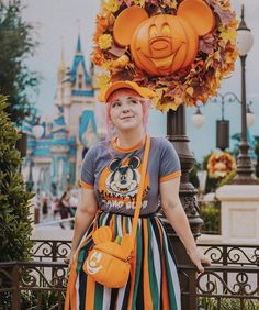 a woman with pink hair wearing a mickey mouse hat and striped dress standing in front of a pumpkin head