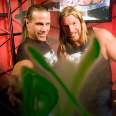 two men standing next to each other in front of a red wall and green plant