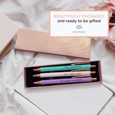 three pens are in a pink box on a white bed with flowers and a notepad