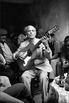 black and white photograph of man playing guitar in front of other people sitting around table