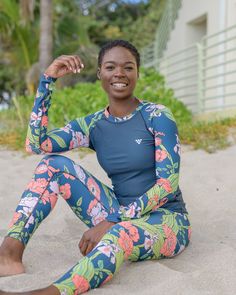 a woman sitting in the sand wearing a blue and pink floral print wetsuit with her arms behind her head