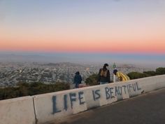 three people standing on the side of a road with graffiti written on it, overlooking a city at sunset