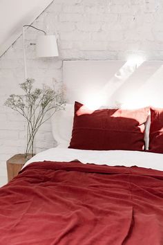 a bed with red sheets and pillows in a white room next to a brick wall