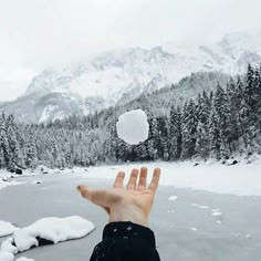 someone is holding their hand out in front of the snow covered mountains and lake with trees