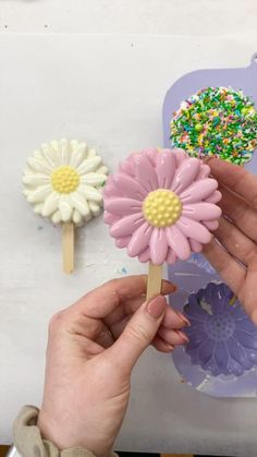 a hand holding a flower shaped object next to two other flowers on a table with sprinkles