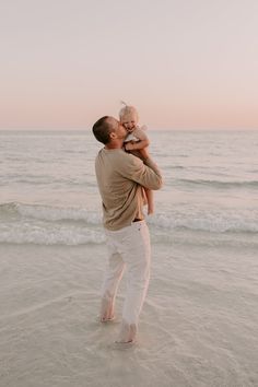 a man holding a baby while standing on top of a beach next to the ocean